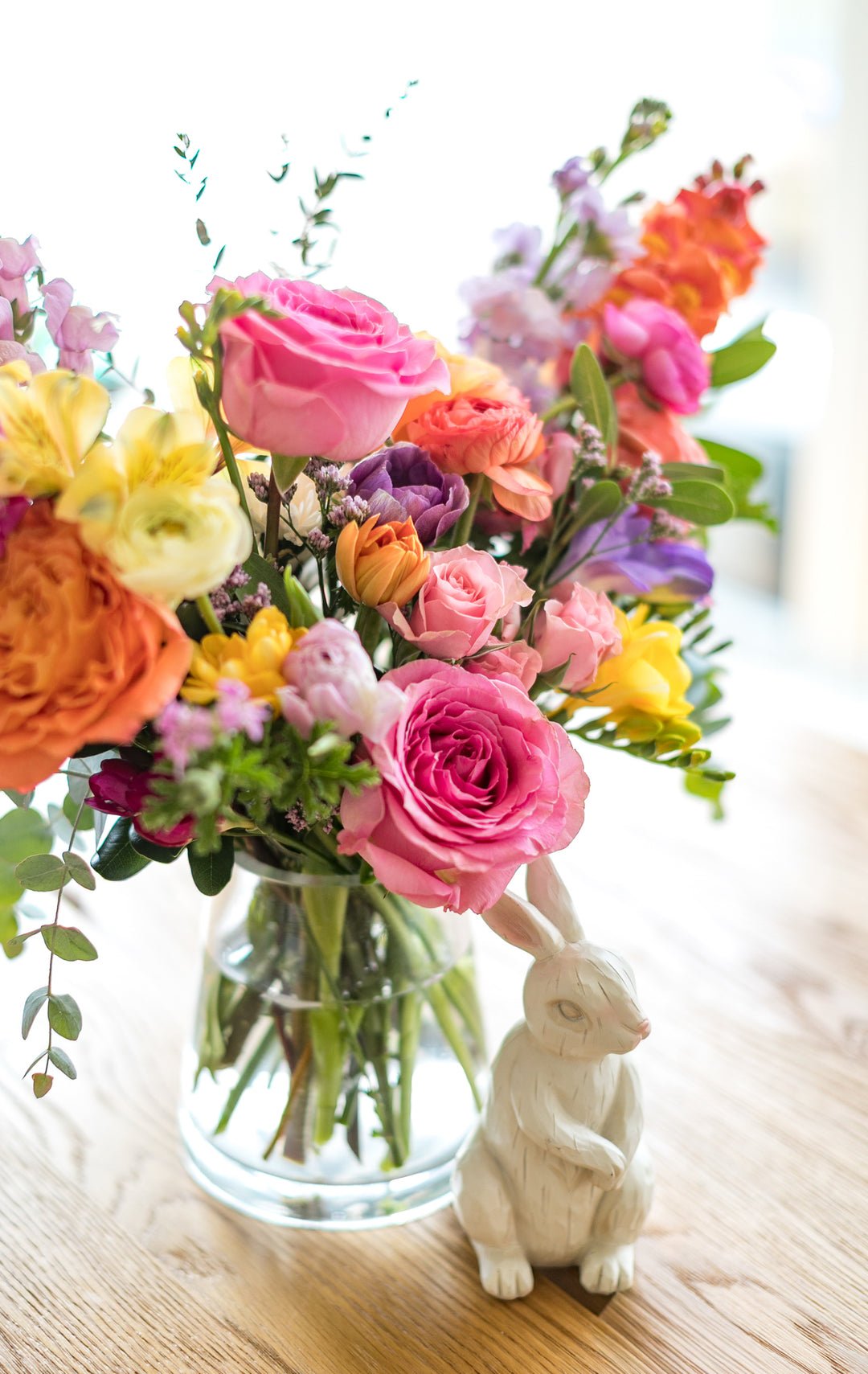 Colorful bouquet of flowers in a clear vase with a white rabbit figurine on a wooden surface.