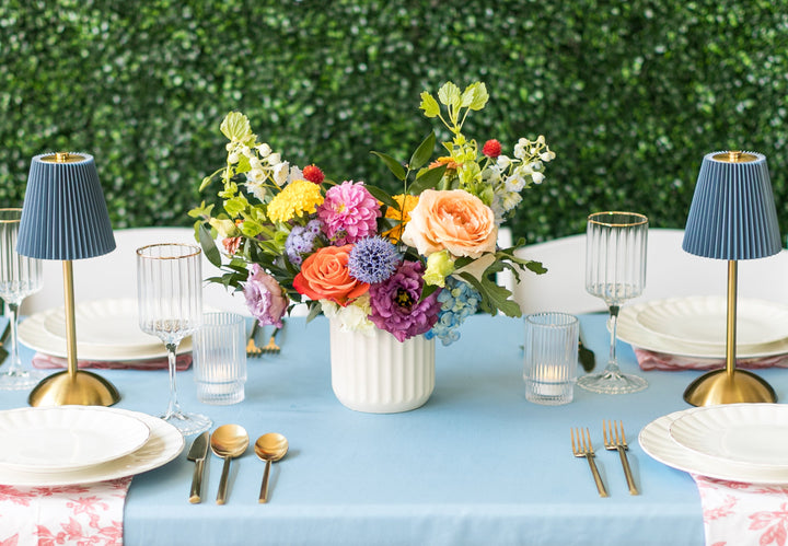 A styled table setting with a light blue tablecloth, white plates, pink floral napkins, gold cutlery, blue lampshades, and a colorful floral centerpiece against a green hedge background. Kansas City wedding florist.