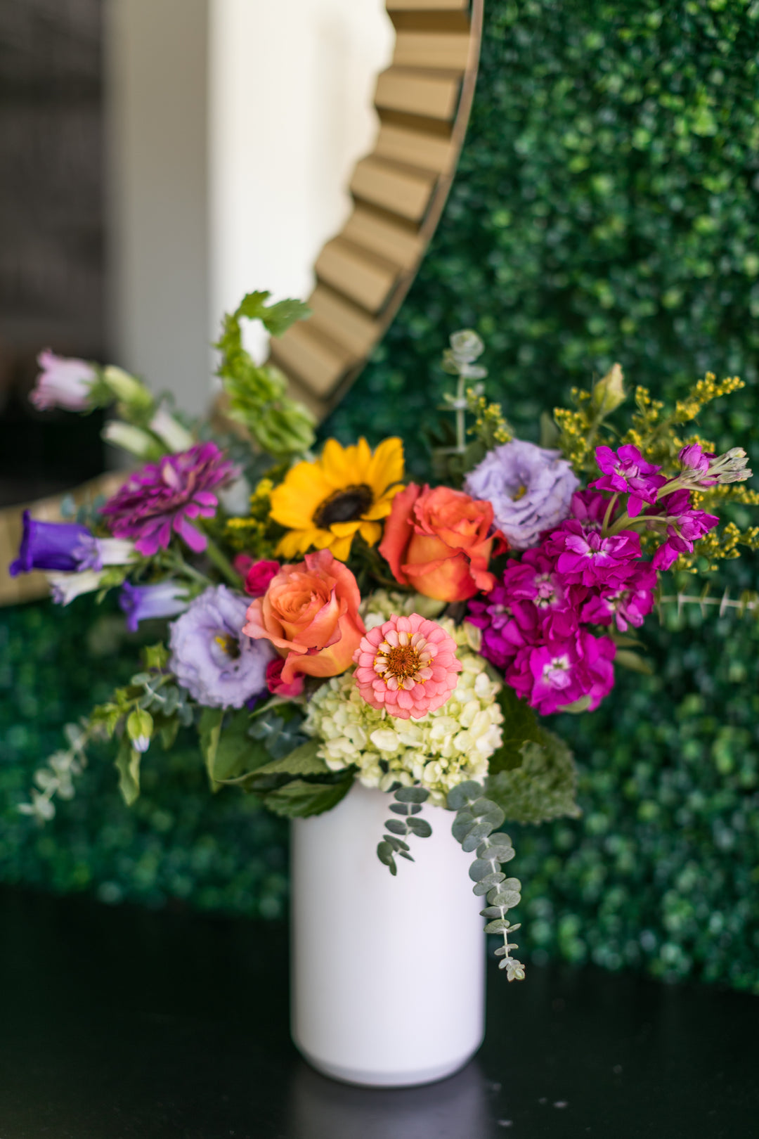 A vibrant, multicolored floral arrangement featuring sunflowers, purple flowers, orange roses, and green foliage in a white ceramic vase, set against a green leaf wall background. All the Colors Bel Fiore Co Downtown Lee's Summit Florist 
