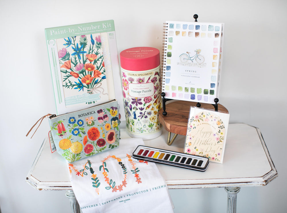 Collection of art supplies including a floral notebook, watercolor set, and coloring book on a white surface.
